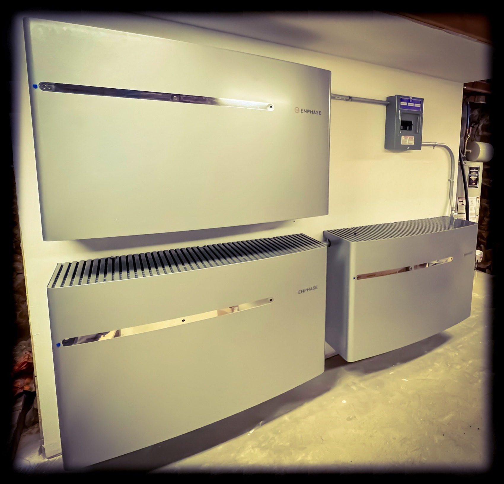 Three wall-mounted battery units beside a subpanel on an unfinished basement wall in a Tri-State home