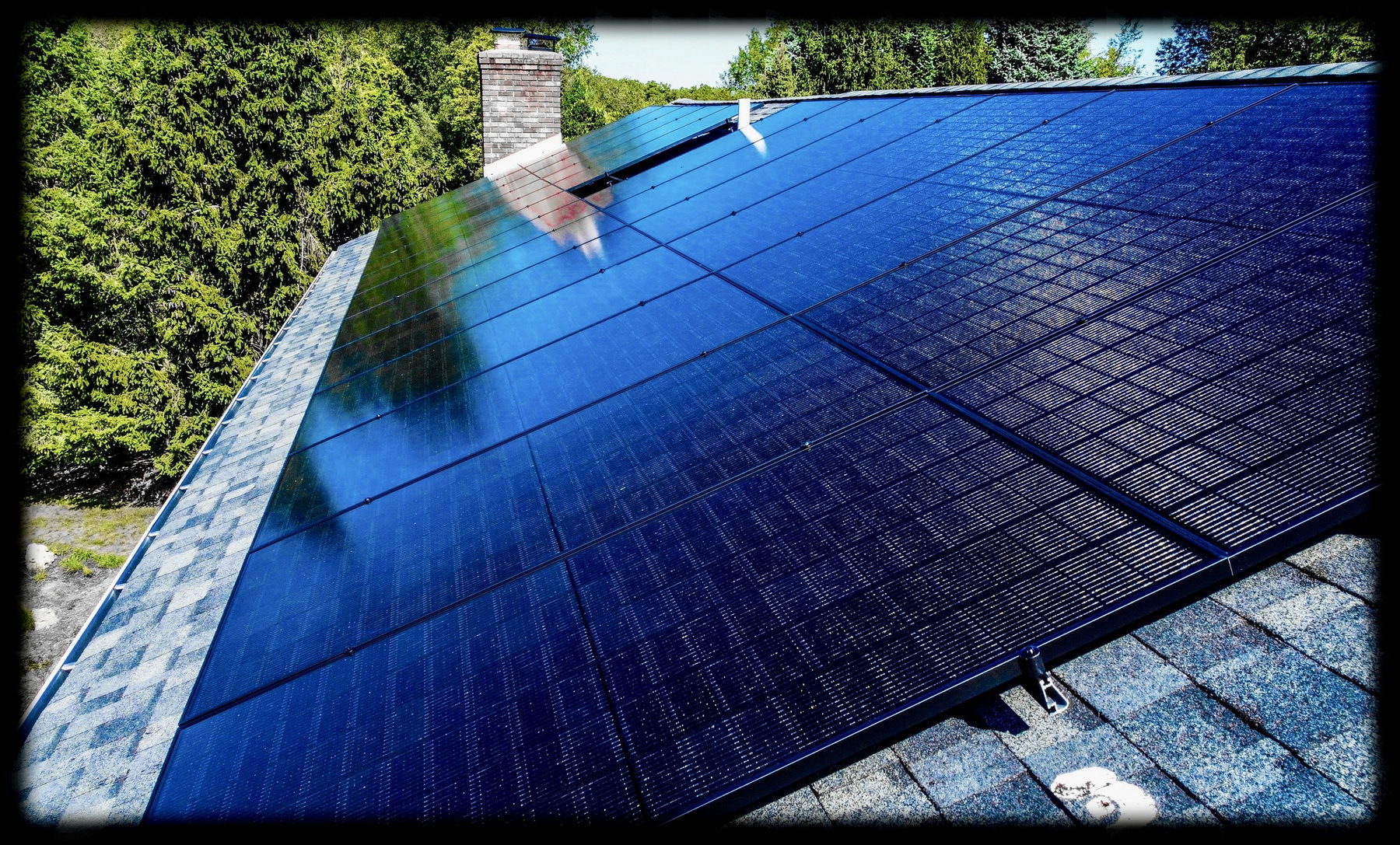 Black all-black solar modules on an asphalt-shingle roof beside a brick chimney, with a treeline behind